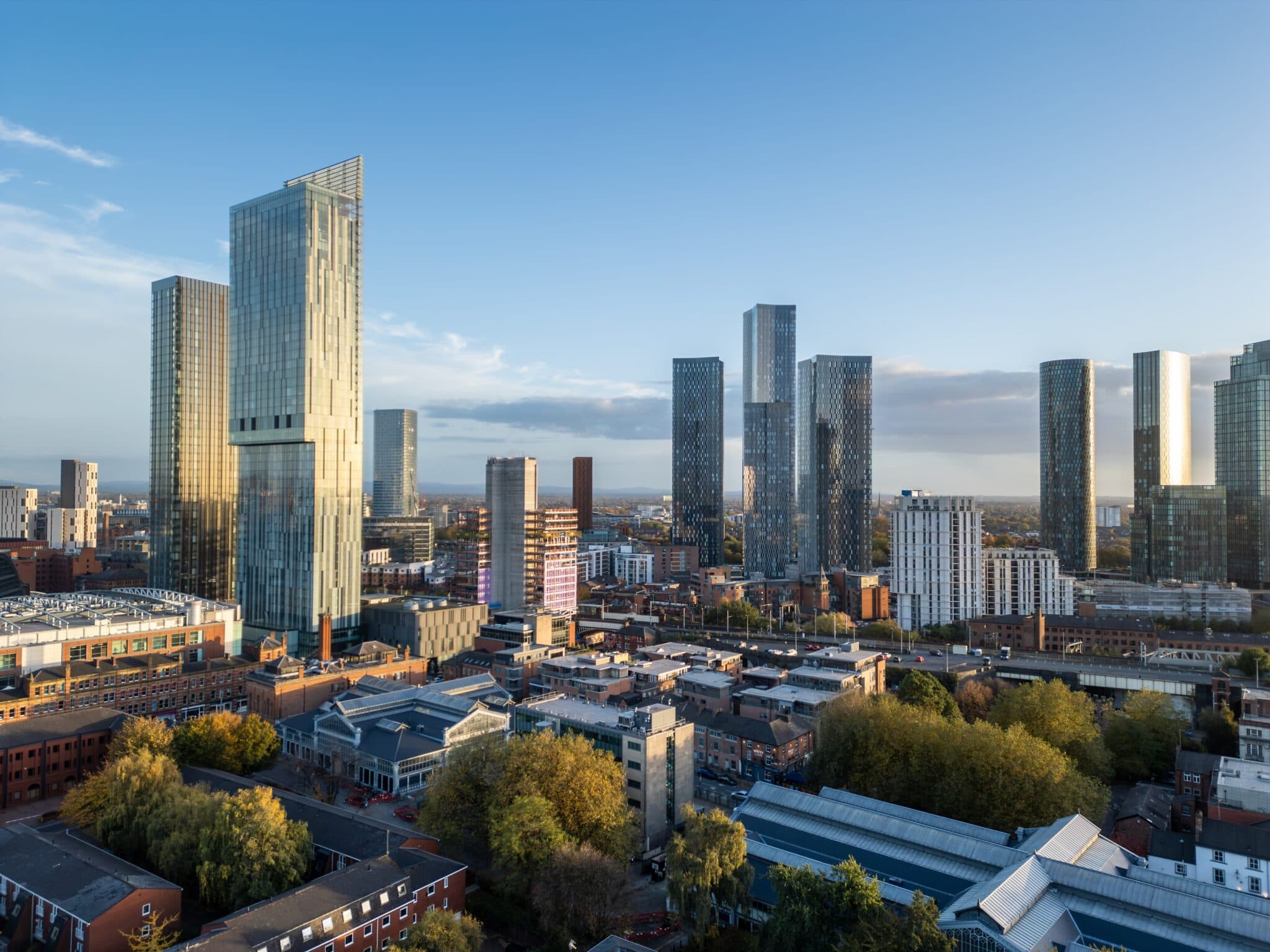 Aerial view of Manchester, England, highlighting the Deansgate area with its mix of modern skyscrapers and historic buildings, surrounded by the wider city skyline in soft October light